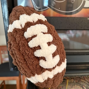 May include: A handmade, brown and white crocheted football. The football is a plush, textured design with white stitching details. The football is a decorative item.