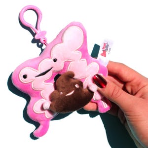May include: A pink plush toy shaped like a stylized colon, with a brown plush poop emoji inside. The toy has a pink clip and a small tag that reads "I Heart Guts! colon".