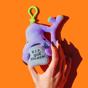 May include: A plush, purple gall bladder toy with green accents and a green clip. The toy has a crying eye and a tombstone that reads "R.I.P. gall bladder". The background is orange.