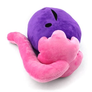 Puede incluir: Un juguete de peluche con forma de gusano morado con una cola rosa. El gusano tiene un ojo negro y una sonrisa simple.
