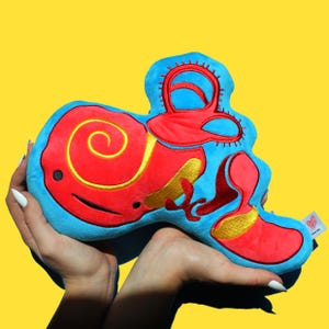 May include: A plush toy shaped like a human ear. The ear is blue with red and yellow details. The toy is being held by two hands.