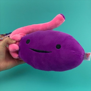 Puede incluir: Juguete de peluche morado con forma de ri&ntilde;&oacute;n con un tubo de peluche rosa adjunto. El ri&ntilde;&oacute;n tiene una cara sonriente con ojos negros y una boca.