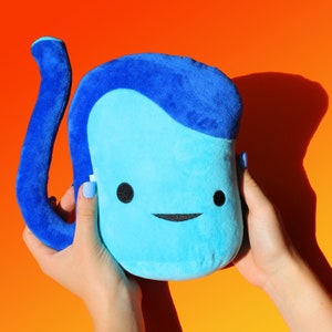 May include: A light blue plush toy with a dark blue curved appendage and a dark blue hair-like design. The toy has black button eyes and a stitched smile. The plush is held against an orange and yellow gradient background.