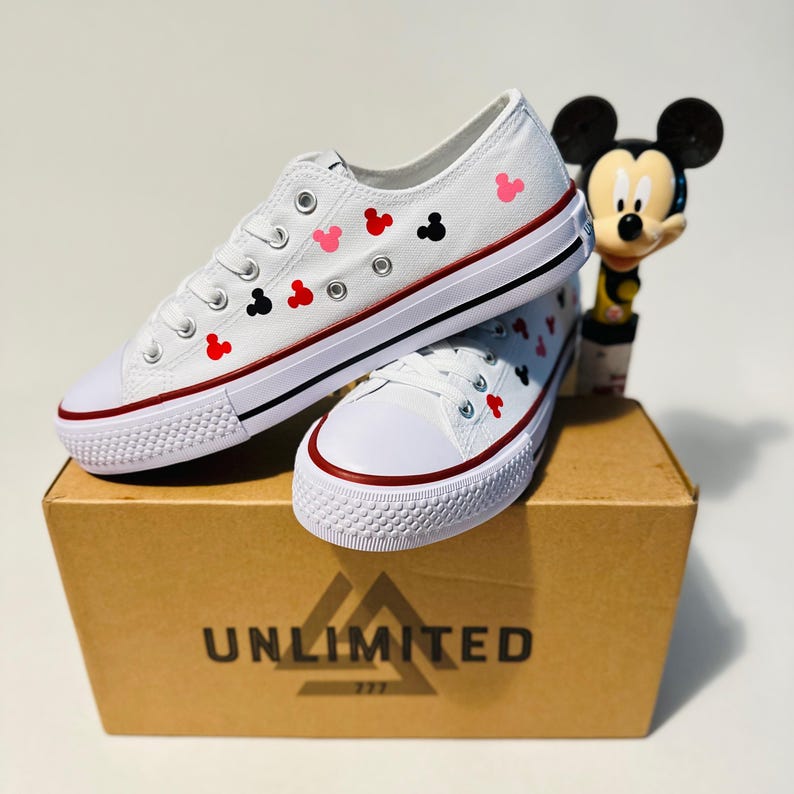 May include: White canvas shoes with red, black and pink Mickey Mouse heads printed on them. The shoes are sitting on a box that says "UNLIMITED 777".