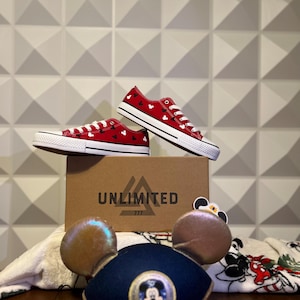 May include: Red canvas shoes with a white Mickey Mouse pattern, sitting on a brown box with the word "UNLIMITED" and the number "777" printed on it. The shoes are next to a white towel with a Mickey Mouse design and a blue Mickey Mouse hat with gold ears.