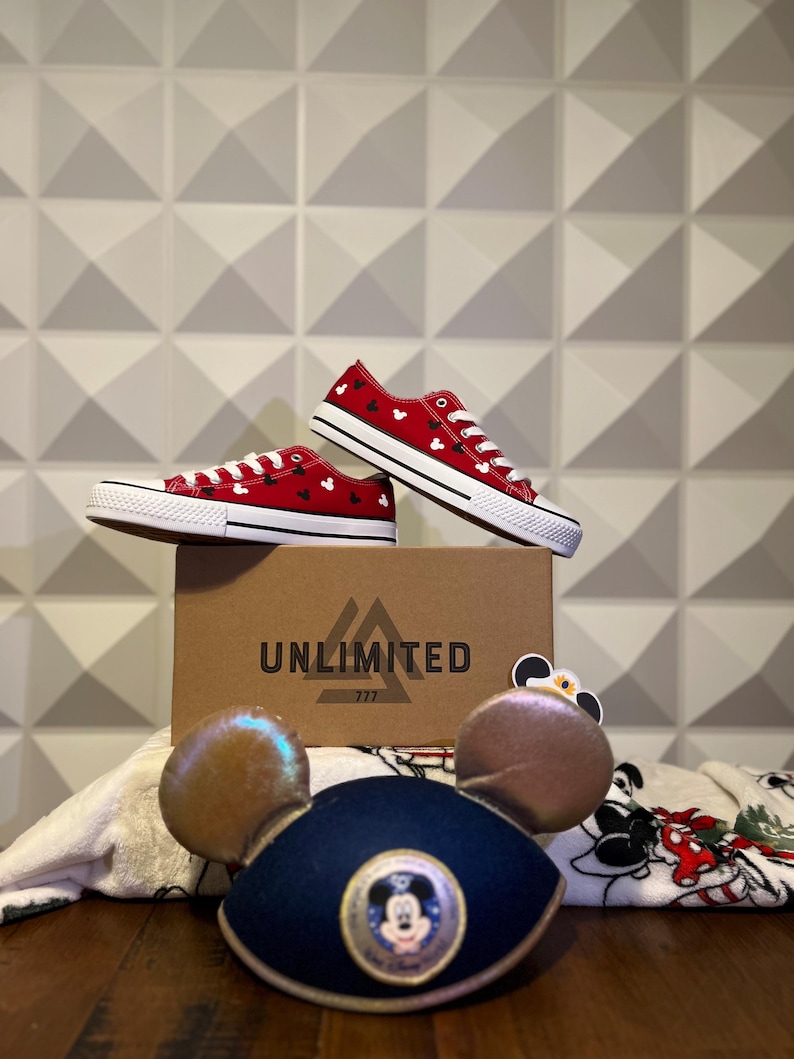 May include: Red canvas shoes with a white Mickey Mouse pattern, sitting on a brown box with the word "UNLIMITED" and the number "777" printed on it. The shoes are next to a white towel with a Mickey Mouse design and a blue Mickey Mouse hat with gold ears.