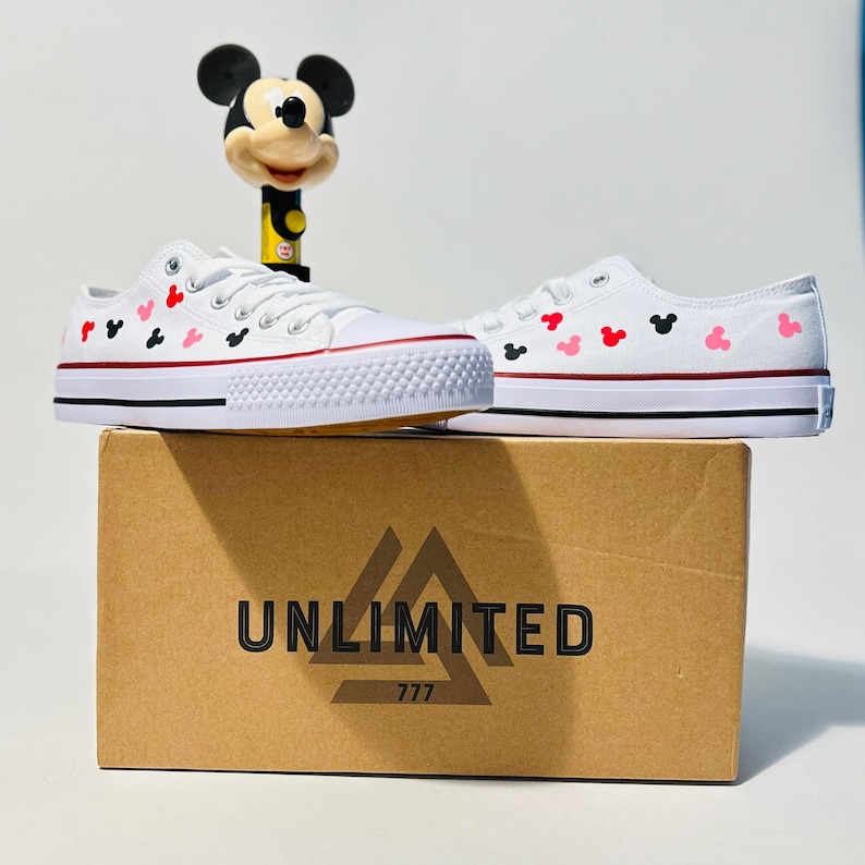 May include: White canvas shoes with red and black Mickey Mouse heads printed on them. The shoes are sitting on a box that says "UNLIMITED 777".