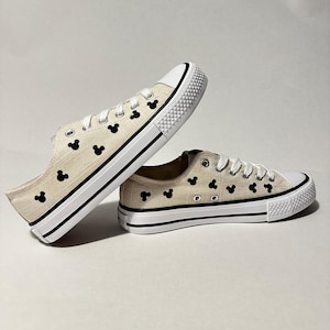 May include: A pair of white canvas trainers with black Mickey Mouse heads printed all over. The shoes have white laces and a black rubber sole.