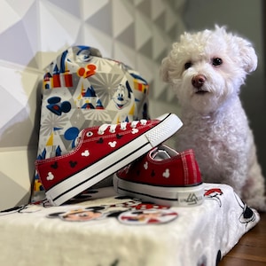 May include: Red canvas sneakers with black Mickey Mouse ears printed on them. The sneakers are sitting on a white blanket with a Mickey Mouse design. A white dog is sitting behind the sneakers.