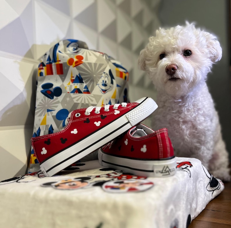 May include: Red canvas sneakers with black Mickey Mouse ears printed on them. The sneakers are sitting on a white blanket with a Mickey Mouse design. A white dog is sitting behind the sneakers.