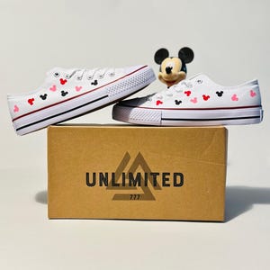 May include: White canvas shoes with red and black Mickey Mouse heads printed on them. The shoes are sitting on a brown box with the word "UNLIMITED" printed on it.