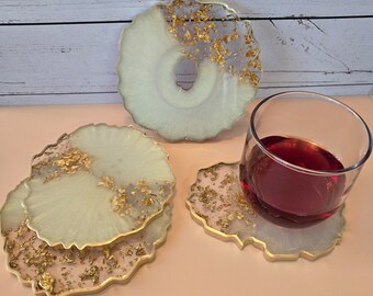 May include: A set of four round, irregular-edged coasters made of translucent white resin with embedded gold leaf flakes and gold-painted rims. One coaster supports a clear glass filled with a dark red liquid, while three others are stacked nearby.