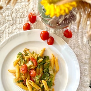 May include: A white plate with a serving of pasta salad with penne pasta, cherry tomatoes, and leafy greens. The plate is on a white tablecloth with a fork and red tomatoes on the side.
