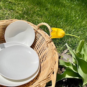May include: Three white ceramic plates with a textured surface are stacked inside a wicker basket. The basket has a handle on the side. Two yellow tulips are in the background.