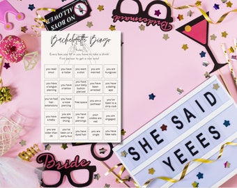 May include: A pink-themed bachelorette party scene with a bingo game, props, and confetti. The bingo card reads "Bachelorette Bingo" with various prompts. Decorative elements include a "She Said Yees" sign, a martini glass, and a "Bride" glasses cutout.