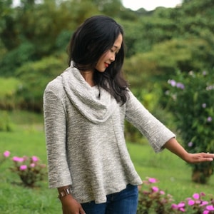 Peut inclure: Une femme portant un pull gris à col roulé avec des manches longues et un motif texturé. Elle se tient dans un champ herbeux avec des fleurs.