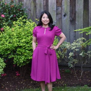 Puede incluir: Una mujer con un vestido magenta con un escote de ojo de cerradura y un lazo en la cintura. Está de pie frente a una valla de madera y arbustos verdes.