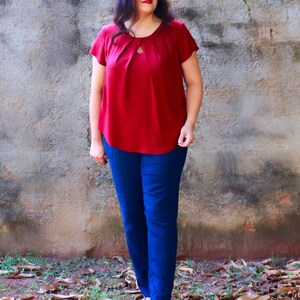 May include: A woman wearing a red top with a keyhole neckline and blue pants. She is standing in front of a stone wall with leaves on the ground.