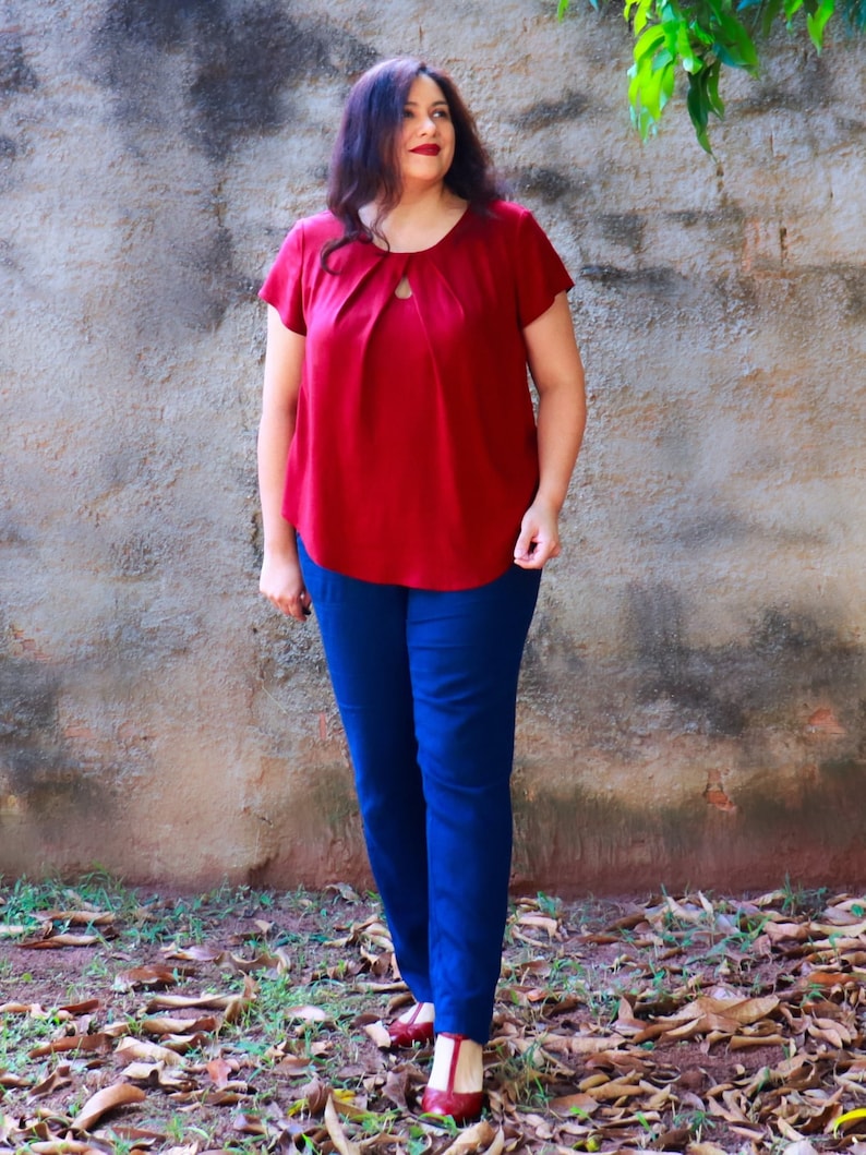 May include: A woman wearing a red top with a keyhole neckline and blue pants. She is standing in front of a stone wall with leaves on the ground.