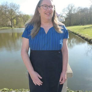 May include: A woman wearing a blue and black striped dress with a V-neckline. She is standing by a pond and looking off to the side.