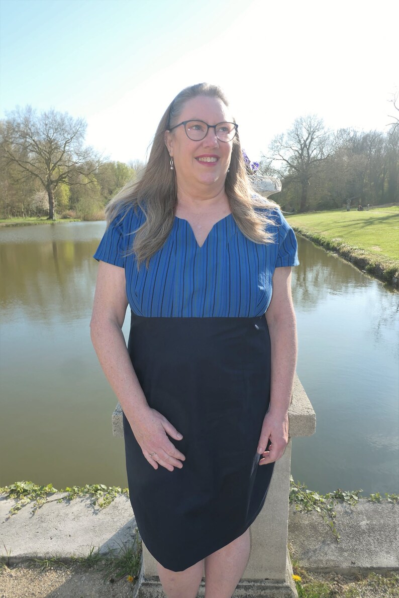 May include: A woman wearing a blue and black striped dress with a V-neckline. She is standing by a pond and looking off to the side.