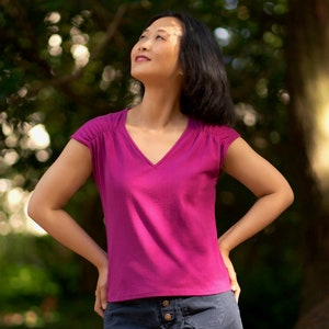 Op de afbeelding: Een vrouw die een magenta V-hals T-shirt met korte mouwen draagt, staand in een park met bomen op de achtergrond.