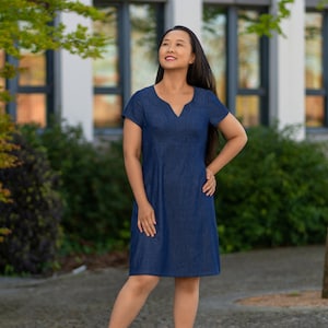 Puede incluir: Una mujer con un vestido de mezclilla azul oscuro con cuello en V y mangas cortas. Está de pie frente a un edificio con ventanas y vegetación verde.