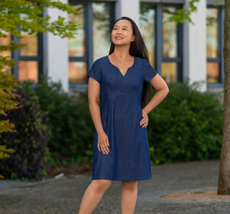 May include: A woman wearing a dark blue denim dress with a v-neck and short sleeves. She is standing in front of a building with windows and green foliage.