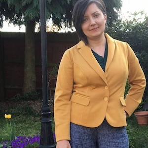 Op de afbeelding: Een mosterdgele blazer met twee knopen en een revers met inkeping. De blazer wordt gedragen over een donkergroene top en gecombineerd met een blauwe broek met patroon. De achtergrond omvat een tuin met een zwarte lantaarnpaal.