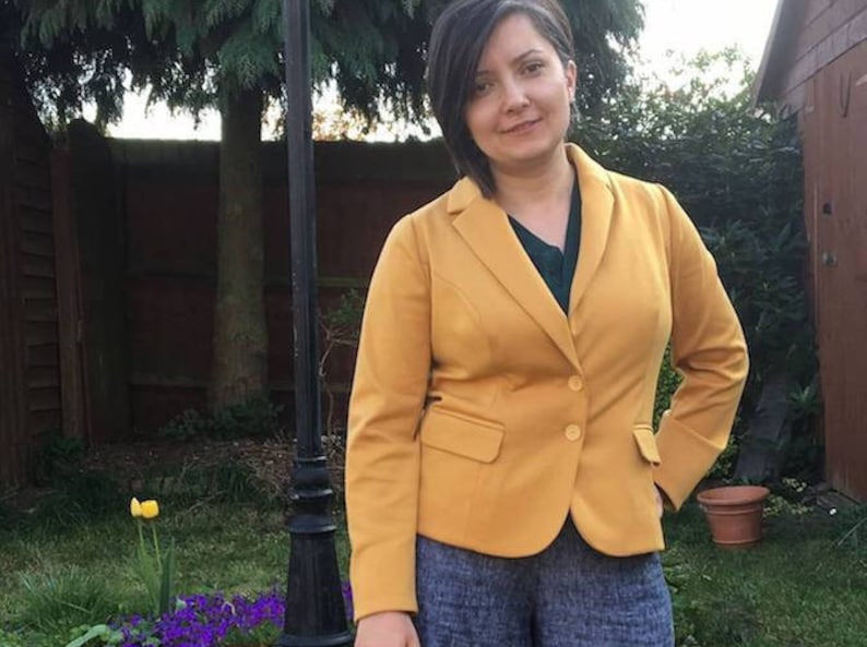 Op de afbeelding: Een mosterdgele blazer met twee knopen en een revers met inkeping. De blazer wordt gedragen over een donkergroene top en gecombineerd met een blauwe broek met patroon. De achtergrond omvat een tuin met een zwarte lantaarnpaal.
