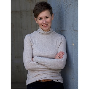 May include: A woman with short brown hair is smiling and wearing a light grey turtleneck sweater with her arms crossed. She is standing against a blue wall.
