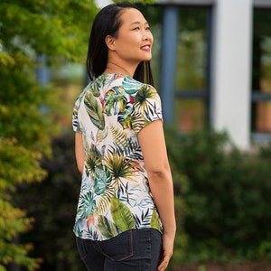 May include: A white blouse with a tropical print featuring green, blue, and yellow leaves. The blouse has a V-neck and short sleeves.