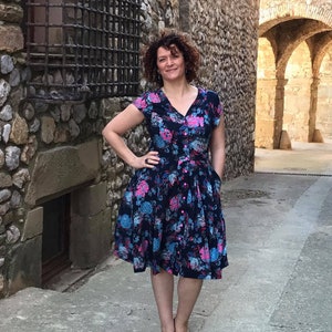 Könnte beinhalten: Eine Frau in einem dunkelblauen Blumenkleid mit rosa und blauen Blumen. Sie steht in einer steingepflasterten Gasse.