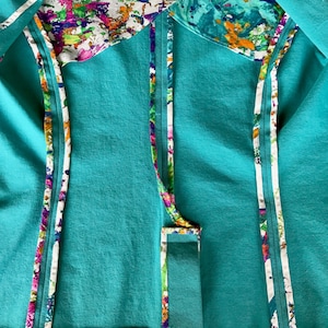 Op de afbeelding: Close-up van een turquoise kledingstuk met kleurrijke abstracte patronen. Het kledingstuk heeft een turquoise buitenkant met witte bies en een levendige binnenvoering. De bies en voering tonen een ontwerp met spetters van roze, paars, groen en oranje.