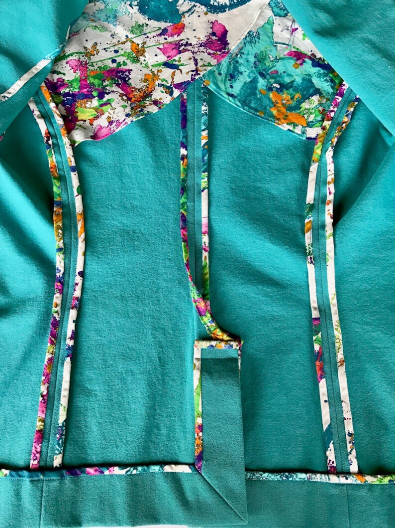 Op de afbeelding: Close-up van een turquoise kledingstuk met kleurrijke abstracte patronen. Het kledingstuk heeft een turquoise buitenkant met witte bies en een levendige binnenvoering. De bies en voering tonen een ontwerp met spetters van roze, paars, groen en oranje.