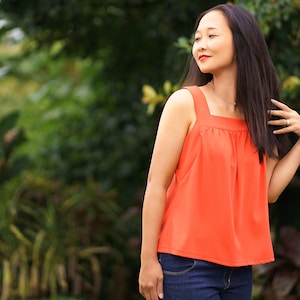 Puede incluir: Una mujer con un top naranja brillante sin mangas, con escote cuadrado y un corpiño fruncido. Está de pie frente a un fondo verde frondoso.