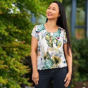 May include: A woman wearing a white blouse with a tropical print and blue jeans. The blouse has a round neckline and short sleeves.