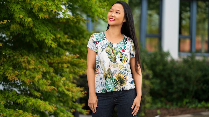 May include: A woman wearing a white blouse with a tropical print and blue jeans. The blouse has a round neckline and short sleeves.