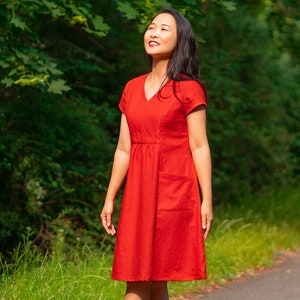 Può includere: Una donna con un vestito rosso si trova in piedi in un'area erbosa con alberi sullo sfondo. Sta guardando il cielo e sorridendo.