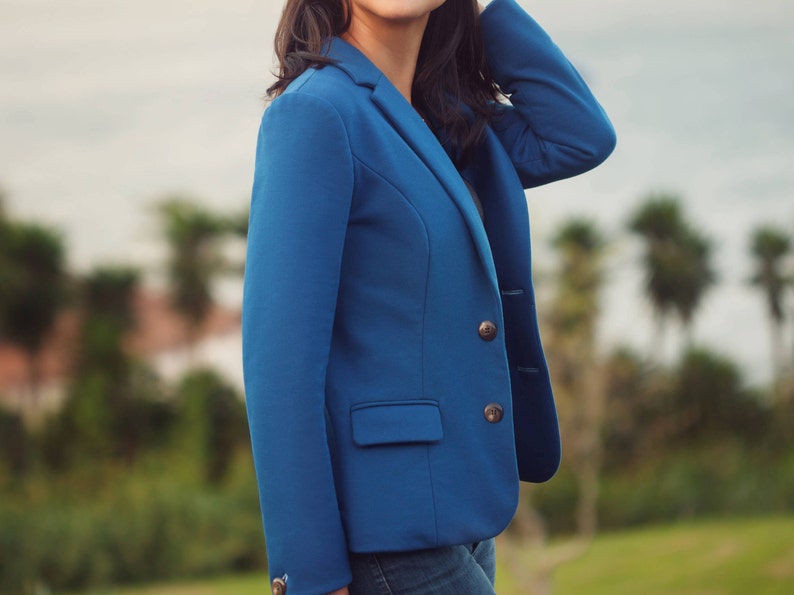 Op de afbeelding: Een levendige blauwe blazer met een klassiek ontwerp. De jas heeft een revers, een sluiting met twee knopen en een enkele borstzak. De blazer heeft twee bruine knopen aan de voorkant en &eacute;&eacute;n op elke mouw. De achtergrond is een vage buitensc&egrave;ne.