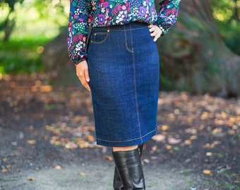 May include: A dark blue denim skirt with a high waist and a straight cut. The skirt has a zipper and button closure in the front and a small pocket on the right side.