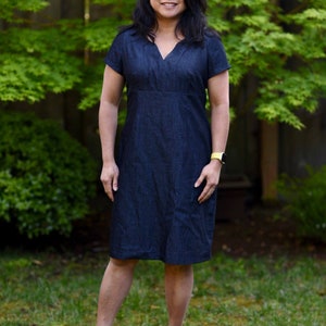 May include: A woman wearing a dark blue denim dress with short sleeves and a V-neck. She is standing in front of a green leafy background.