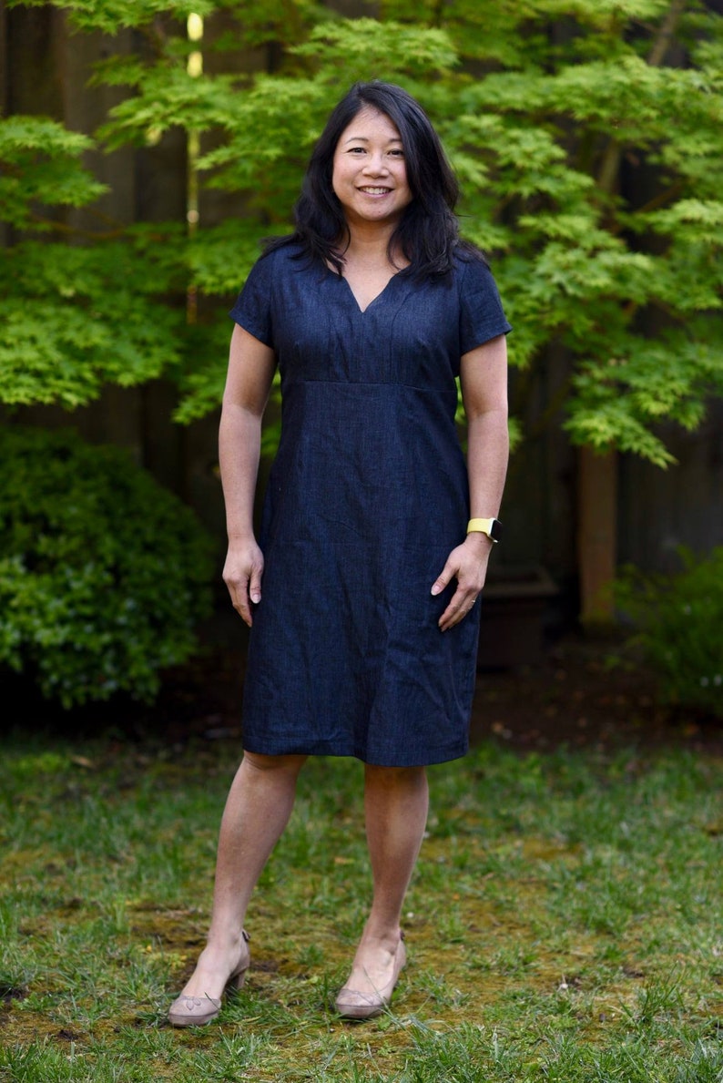 May include: A woman wearing a dark blue denim dress with short sleeves and a V-neck. She is standing in front of a green leafy background.
