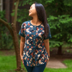 May include: A woman wearing a teal blue blouse with a floral print. The blouse has a button-up front and short sleeves. The woman is standing in a grassy area with trees in the background.