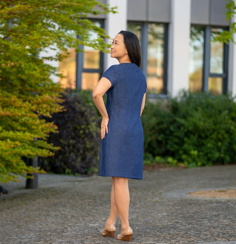 May include: A woman wearing a dark blue denim dress with short sleeves and a button-up front. She is standing outdoors with her back to the camera and looking over her shoulder.