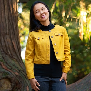 May include: A woman with long black hair is wearing a yellow denim jacket, a black mock neck shirt, and blue jeans. She is standing in front of a tree with a blurred background of green foliage.