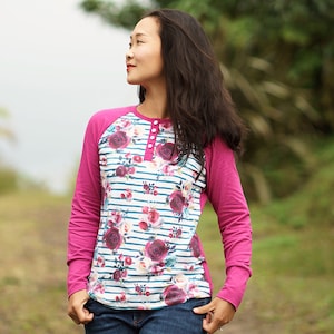 May include: A woman wearing a long-sleeved shirt with a floral and striped pattern. The shirt has a pink raglan sleeve design and a single button closure at the neckline.
