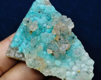 Puede incluir: Primer plano de un espécimen mineral con un vibrante tono turquesa. La superficie está cubierta de pequeñas formaciones redondeadas, con grupos de esferas translúcidas de color melocotón pálido. La base del espécimen tiene una textura rugosa, gris-marrón.