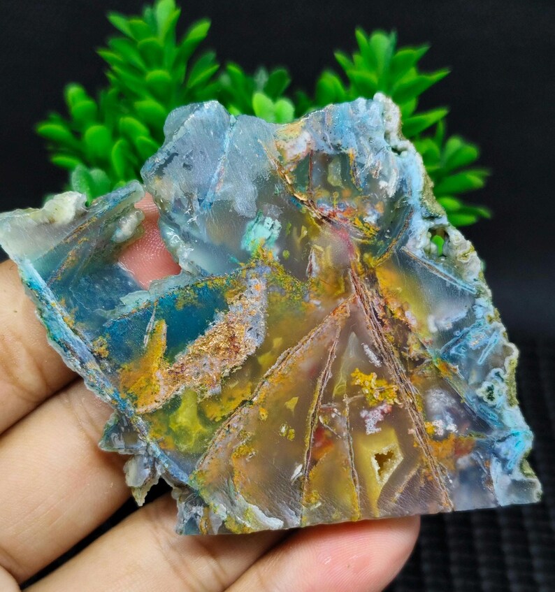 May include: A slice of agate with a variety of colors, including blue, green, yellow, and brown. The agate has a natural, rough texture and is translucent.
