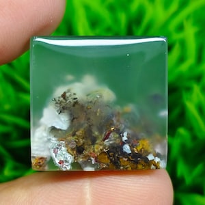 以下が含まれることがあります： 茶色、白、銀色のインクルージョンが入った正方形の半透明な緑色のアゲートカボション。アゲートは透明なレジンにセットされています。